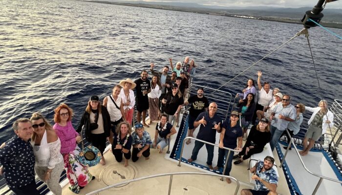 A large group of adults poses and celebrates together on the deck of a Blue Dolphin Kauaʻi catamaran at sunset, with the open Pacific Ocean and a distant coastline visible in the background, crew members visible among the guests.