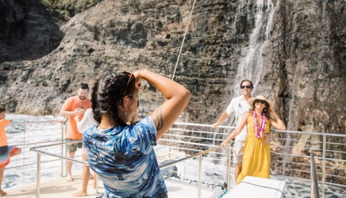 A Blue Dolphin Kauaʻi crew member in a blue aloha shirt photographs a smiling couple posing on the bow of a catamaran in front of a waterfall streaming down dark lava rock cliffs of the Nāpali Coast, with other guests visible on deck.